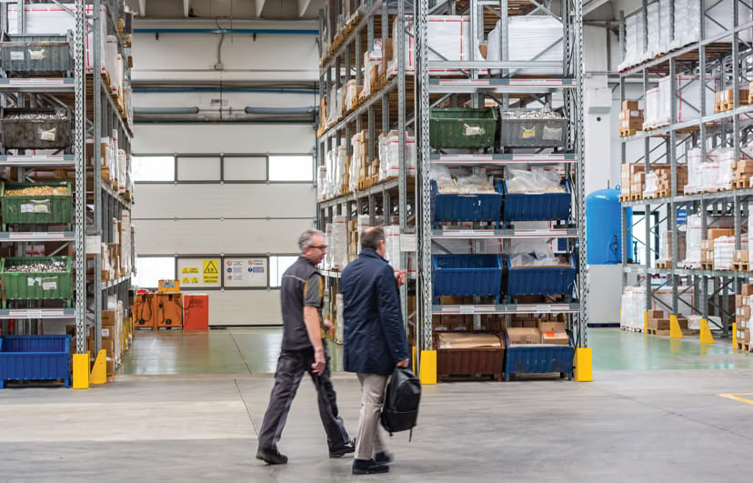 Two people walking through the warehouse past shelves of packed parts